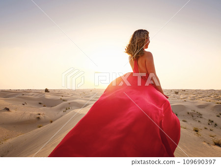 Desert adventure. Young arabian Woman in red silk dress in sands dunes of UAE desert at sunset, fantastic view. The Dubai Desert Conservation Reserve, United Arab Emirates. 109690397