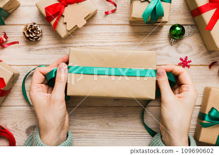 Woman is packing surprise gift for Christmas holidays, sitting at workplace with craft tools, top view. Woman wrapping Christmas gift at colored table 109690602