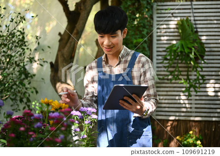 Smiling male floral shop owner wearing apron using digital tablet and checking stock Smiling male floral shop owner wearing apron using digital tablet and checking stock 109691219