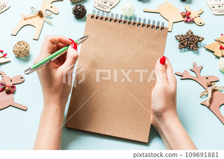 Top view of female hand making some notes in noteebok on blue background. New Year decorations and toys. Christmas time concept Top view of female hand making some notes in noteebok on blue background. New Year decorations and toys. Christmas time concept 109691881