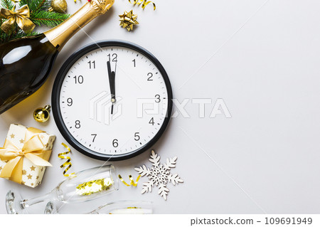 New year celebration concept with a bottle of champagne and two glasses toasting. Christmas gift box, alarm clock and fir tree branch on colored table. Top view Copy space 109691949