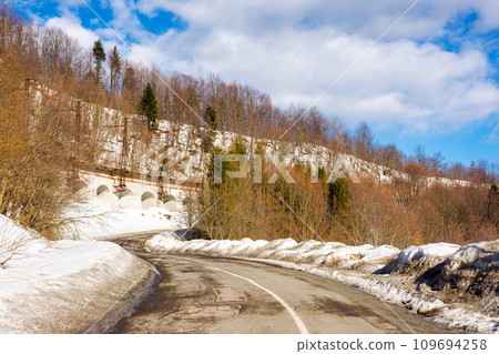 road pass winding through forested snow covered hills. mountainous winter landscape on a sunny day beneath a blue sky with clouds 109694258