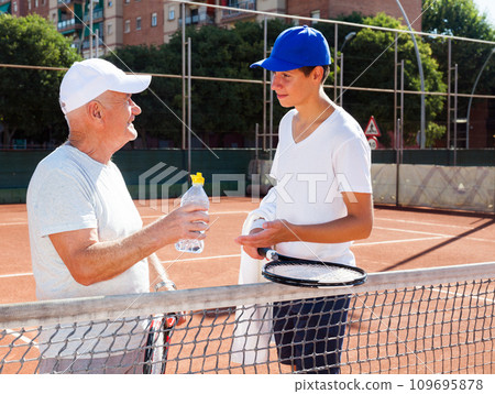 Grandfather and grandson talking on court playing tennis Grandfather and grandson talking on court playing tennis 109695878
