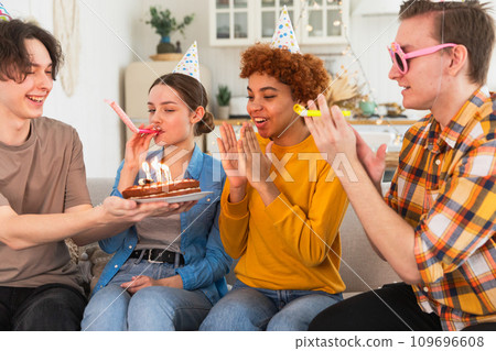 Make a wish. Woman wearing party cap blowing out burning candles on birthday cake. Happy Birthday party. Group of friends wishes girl happy birthday. People celebrating birthday with party at home Make a wish. Woman wearing party cap blowing out burning candles on birthday cake. Happy Birthday party. Group of friends wishes girl happy birthday. People celebrating birthday with party at home 109696608