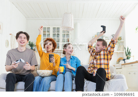 Home party. Cheerful group of friends playing video games at home. Happy diverse group buddies having fun together indoor. Friendship leisure entertainment concept. Young best friends enjoying weekend Home party. Cheerful group of friends playing video games at home. Happy diverse group buddies having fun together indoor. Friendship leisure entertainment concept. Young best friends enjoying weekend 109696691