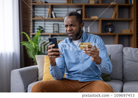 Upset and sad man sitting at home on couch in living room, cheated senior mature african american man displeased holding phone and bank credit card, rejected fund transfer error, account block. 109697453