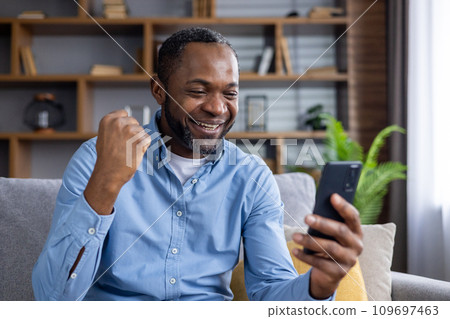 Online winner man sitting at home on sofa in living room, happy successful african american man celebrating victory, holding hand up, received online notification on phone, happy and satisfied. 109697463