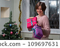 A woman with a pile of Christmas gifts in her hands stands next to a decorated Christmas tree 109697631