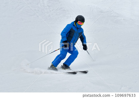A man in blue sportswear on skis rides down a snowy slope A man in blue sportswear on skis rides down a snowy slope 109697633