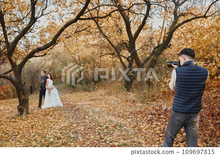 Professional wedding photographer taking pictures of the bride and groom in nature in autumn Professional wedding photographer taking pictures of the bride and groom in nature in autumn 109697815