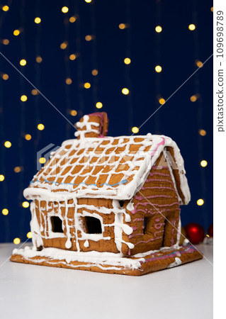 Christmas house made of gingerbread cookies decorated with sugar icing on blue background with bokeh garland Christmas house made of gingerbread cookies decorated with sugar icing on blue background with bokeh garland 109698789