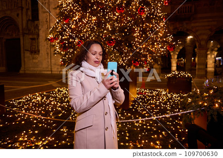 Beautiful woman holds smart mobile phone, standing on the illuminated square with beautiful Christmas tree and garlands Beautiful woman holds smart mobile phone, standing on the illuminated square with beautiful Christmas tree and garlands 109700630
