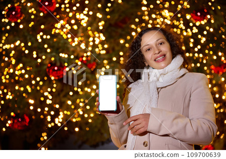 Cheerful Latina woman showing mobile phone with white mockup screen, for ads or application, against lighted Xmas tree 109700639