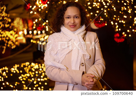 Beautiful woman in warm clothes, smiling looking at camera, standing against illuminated street background 109700640