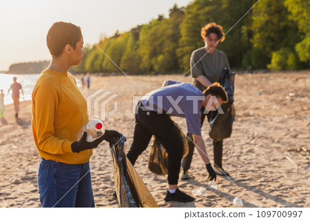 Earth day. Volunteers activists team collects garbage cleaning of beach coastal zone. Woman puts plastic bottle trash in garbage bag on ocean shore. Environmental conservation coastal zone cleaning 109700997