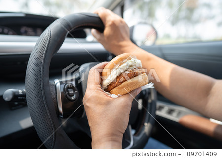 Asian lady holding hamburger to eat in car, dangerous and risk an accident. Asian lady holding hamburger to eat in car, dangerous and risk an accident. 109701409