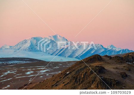 Soft pink light glides over the snow-capped peaks of the mountai 109703029