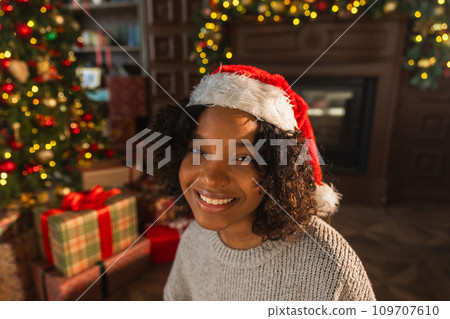 Merry Christmas. African American woman smiling near Christmas tree in classical dark interior. Happy girl in Santa hat in living room with fireplace Christmas tree gift boxes. Christmas eve at home 109707610