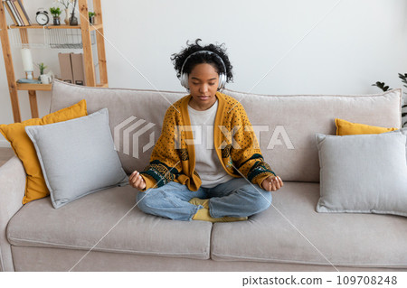 Yoga mindfulness meditation. Young healthy african girl practicing yoga at home. Woman sitting in lotus pose on couch meditating smiling relaxing indoor. Girl doing breathing practice. Yoga at home Yoga mindfulness meditation. Young healthy african girl practicing yoga at home. Woman sitting in lotus pose on couch meditating smiling relaxing indoor. Girl doing breathing practice. Yoga at home 109708248