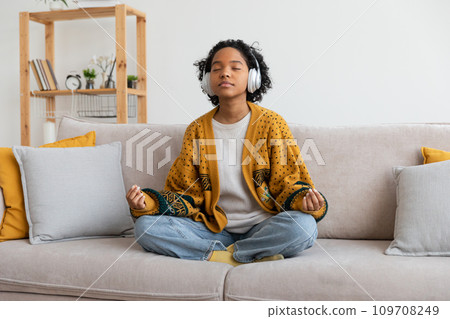 Yoga mindfulness meditation. Young healthy african girl practicing yoga at home. Woman sitting in lotus pose on couch meditating smiling relaxing indoor. Girl doing breathing practice. Yoga at home 109708249