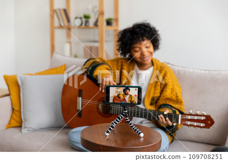 Blogger guitarist. African american girl blogger playing guitar talking to webcam recording vlog. Social media influencer woman streaming at home indoors. Music content creator broadcast tutorial Blogger guitarist. African american girl blogger playing guitar talking to webcam recording vlog. Social media influencer woman streaming at home indoors. Music content creator broadcast tutorial 109708251