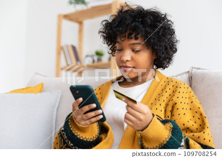 African american woman shopping online holding smartphone paying with gold credit card. Girl sitting at home buying on Internet enter credit card details. Online shopping ecommerce delivery service 109708254