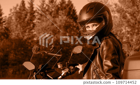 Biker girl riding touring motorbike on forest road. Image toned in Peach Fuzz color of the year 2024. Beautiful woman with red hair in leather jacket sitting on motorcycle in the park. 109714452