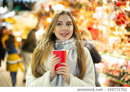 Pretty girl walking in colorful Christmas street market, holding paper cup of warm drink 109715702