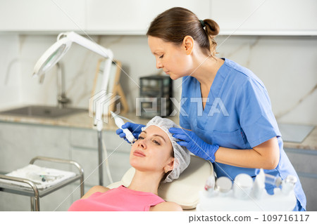 Adult woman cosmetologist doing microcurrent procedure to young woman 109716117