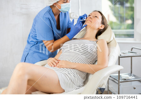 Doctor in disposable mask performs procedure of lip augmentation of girl patient in clinic. 109716441