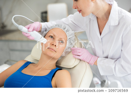 Woman lying on couch during cosmetological ultrasonic facial cleansing in clinic. Professional moisturizing and rejuvenating hardware procedures for people Woman lying on couch during cosmetological ultrasonic facial cleansing in clinic. Professional moisturizing and rejuvenating hardware procedures for people 109716569