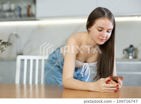 Positive smiling young girl leaned on table and holds smartphone in hands reads news 109716747