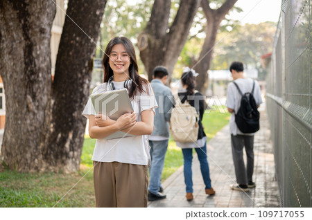 A positive, cute Asian female student with her laptop is standing on a footpath on her campus. 109717055