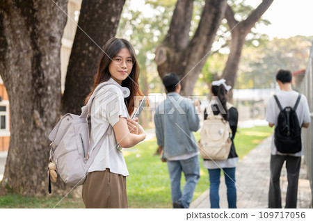 A positive, cute Asian female student with her laptop is standing on a footpath on her campus. 109717056