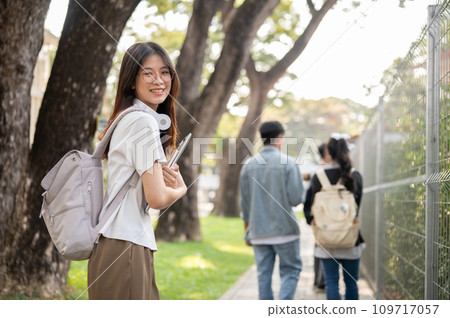A positive, cute Asian female student with her laptop is standing on a footpath on her campus. 109717057