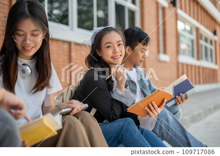 A positive, cute young Asian female college student sits on a footpath with her friends. A positive, cute young Asian female college student sits on a footpath with her friends. 109717086