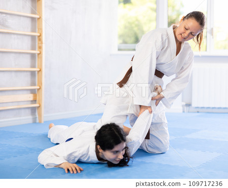 Sportive woman holds her opponent by force while sitting on mats during judo classes Sportive woman holds her opponent by force while sitting on mats during judo classes 109717236