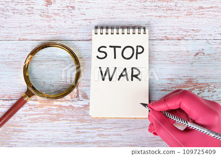 STOP WAR writing on a notebook lying on old boards next to a magnifying glass and a hand in a red medical glove with a pencil STOP WAR writing on a notebook lying on old boards next to a magnifying glass and a hand in a red medical glove with a pencil 109725409