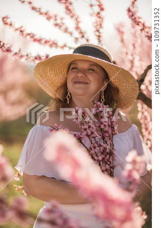 Woman blooming peach orchard. Against the backdrop of a picturesque peach orchard, a woman in a long white dress and hat enjoys a peaceful walk in the park, surrounded by the beauty of nature. 109731352