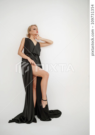 Woman in black dress on white background in the studio. Elegant lady in long gown showing leg. Sexy girl in formal high slit prom dress. Woman in black dress on white background in the studio. Elegant lady in long gown showing leg. Sexy girl in formal high slit prom dress. 109731354