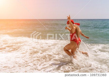 Women in Santa Claus hats run into the sea dressed in red swimsuits. Celebrating the New Year in a hot country 109731360