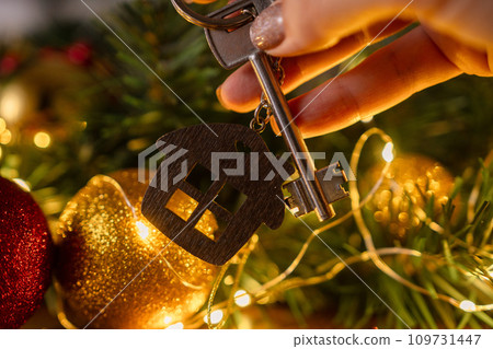 Gift key house with a keychain in hand backdrop of Christmas tree. Building, design, project, moving to new house, mortgage, rent and purchase real estate. A gift for New Year, Christmas. 109731447