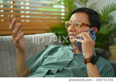 Smiling middle age man having pleasant phone conversation while relaxing on couch at home. Smiling middle age man having pleasant phone conversation while relaxing on couch at home. 109732356