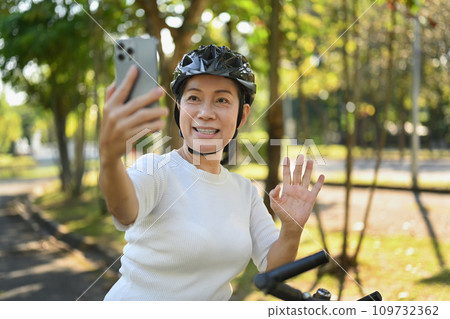 Cheerful middle age woman having video call on mobile phone while resting from cycling in a green park. Cheerful middle age woman having video call on mobile phone while resting from cycling in a green park. 109732362