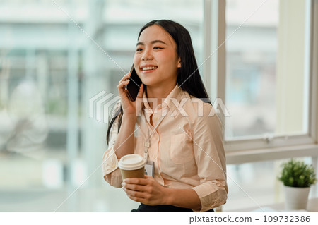 Cheerful Asian female employee talking on mobile phone near office window. 109732386