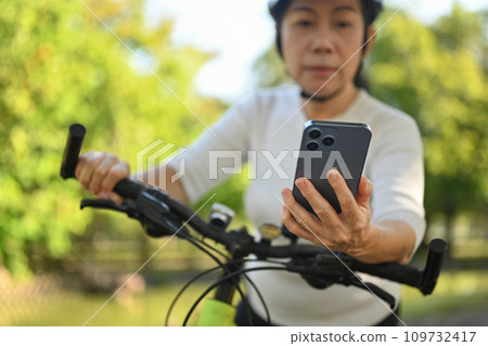 Senior woman hand using mobile phone checking route map location while riding exercise bicycle. Senior woman hand using mobile phone checking route map location while riding exercise bicycle. 109732417