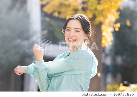 Autumn/winter of a young woman exercising in the autumn leaves, stretching during warm-up exercises, looking at the camera Autumn/winter of a young woman exercising in the autumn leaves, stretching during warm-up exercises, looking at the camera 109733650