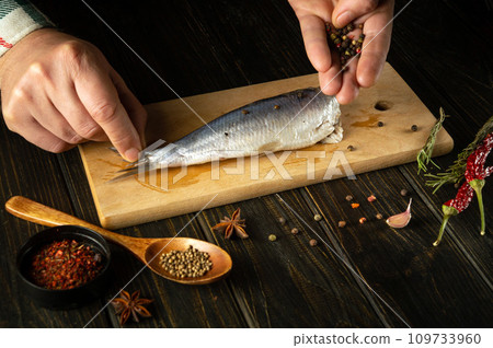 Cooking pickled herring with aromatic spices by the hands of a cook on the kitchen table. Fish diet concept on dark background Cooking pickled herring with aromatic spices by the hands of a cook on the kitchen table. Fish diet concept on dark background 109733960