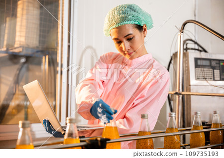 A female engineer oversees beverage bottles on a conveyor belt in the factory. Employing a laptop she guarantees liquid quality adhering to industry standards. 109735193