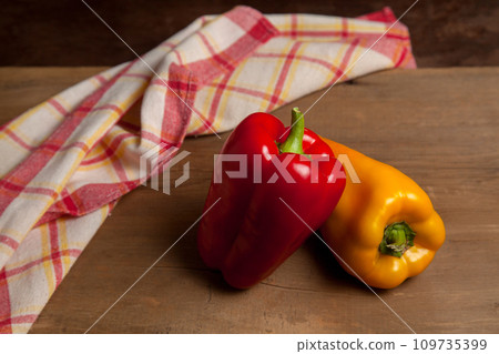 Two red and yellow bell pepper on wooden background with red kitchen towel. . Two red and yellow bell pepper on wooden background with red kitchen towel. . 109735399